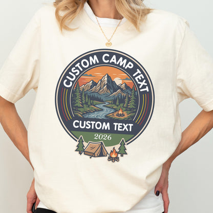 May include: A cream-colored t-shirt featuring a circular graphic with a mountain landscape, a river, and a campfire. The text reads CUSTOM CAMP TEXT at the top and bottom, with 2026 below. A small tent and trees are depicted at the base of the design.