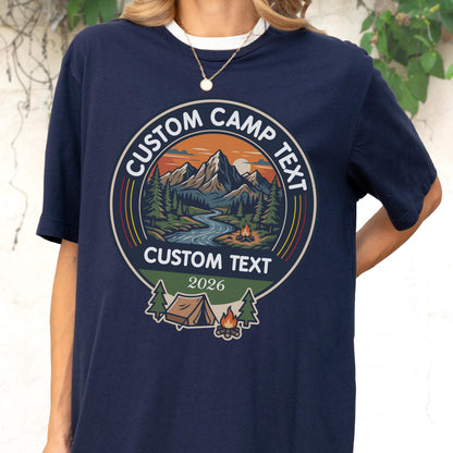 May include: A navy blue t-shirt featuring a circular graphic with a mountain landscape, a river, and a campfire. The text reads CUSTOM CAMP TEXT, CUSTOM TEXT, and 2026. Below the circle is a tent, a campfire, and two pine trees in a flat design.