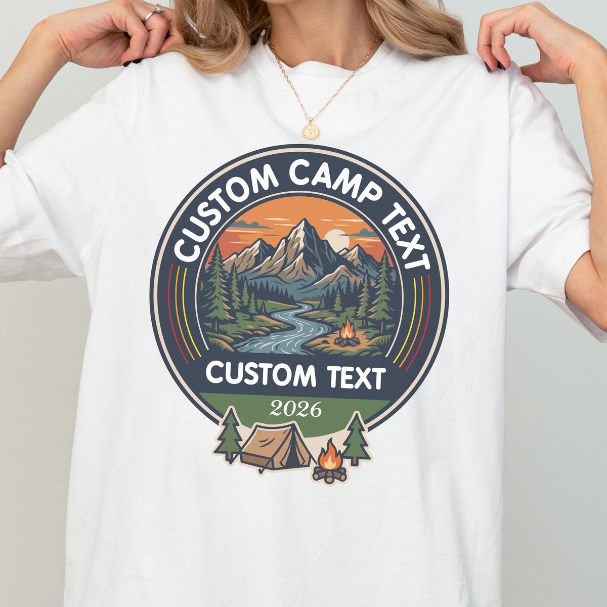 May include: A white t-shirt featuring a circular graphic with a mountain landscape, a river, pine trees, a tent, and a campfire. The text reads CUSTOM CAMP TEXT at the top, CUSTOM TEXT in the center, and 2026 at the bottom of the circular design.