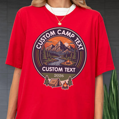 May include: A bright red short-sleeved t-shirt featuring a circular graphic with a mountain landscape, a river, a campfire, and a tent. The text reads CUSTOM CAMP TEXT at the top and bottom, with the year 2026 printed in the center of the design.
