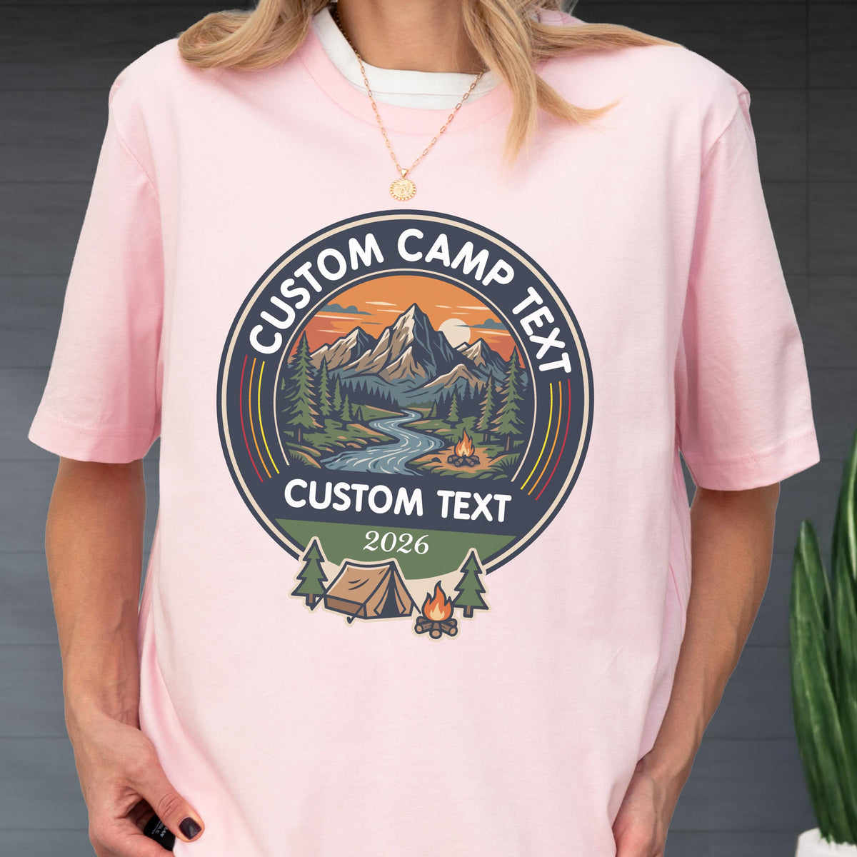 May include: A light pink short-sleeve t-shirt featuring a circular graphic with a mountain landscape, a river, and a campfire. The text reads CUSTOM CAMP TEXT at the top and bottom, with 2026 below, plus a small tent and campfire icon below.
