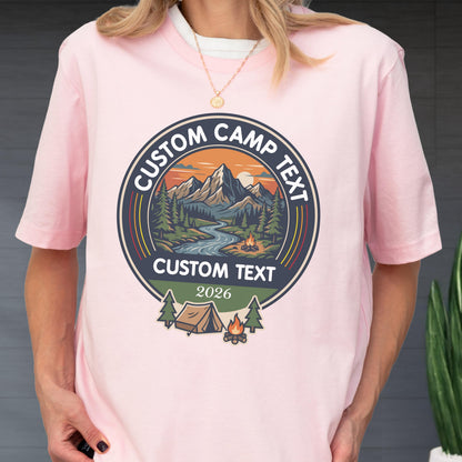 May include: A light pink short-sleeve t-shirt featuring a circular graphic with a mountain landscape, a river, and a campfire. The text reads CUSTOM CAMP TEXT at the top and bottom, with 2026 below, plus a small tent and campfire icon below.