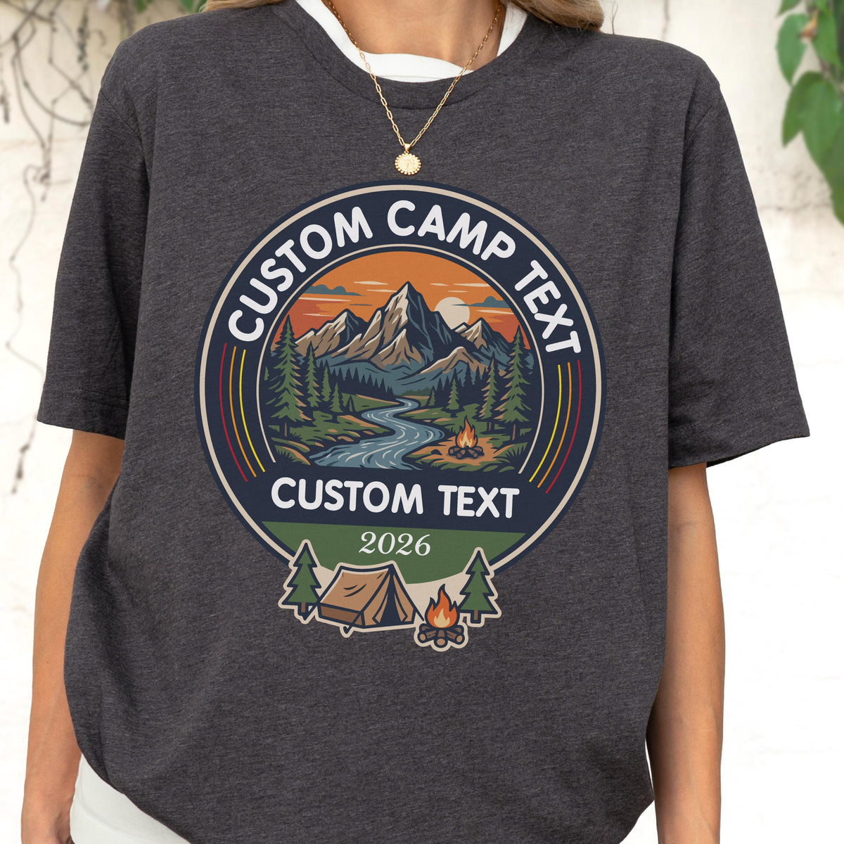 May include: A charcoal gray t-shirt featuring a circular graphic with a mountain landscape, a river, and a campfire. The text reads CUSTOM CAMP TEXT at the top and bottom, with the year 2026 below. A small tent and pine trees are at the base.