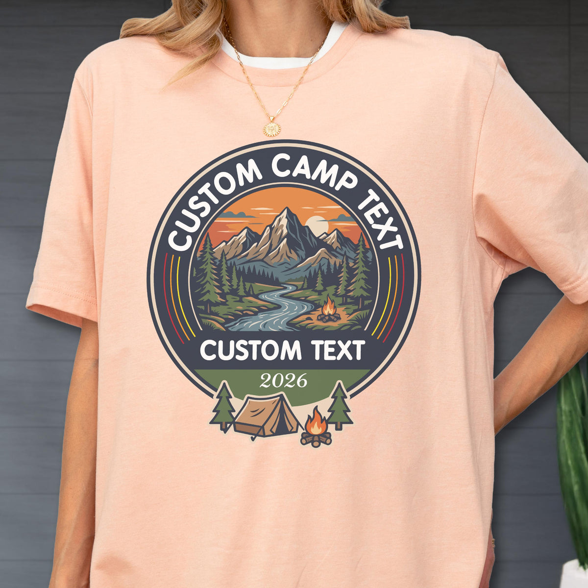 May include: A peach-colored t-shirt featuring a circular graphic with a mountain landscape, a river, pine trees, and a campfire. The text reads CUSTOM CAMP TEXT at the top, CUSTOM TEXT in the center, and 2026 at the bottom, with a tent icon.