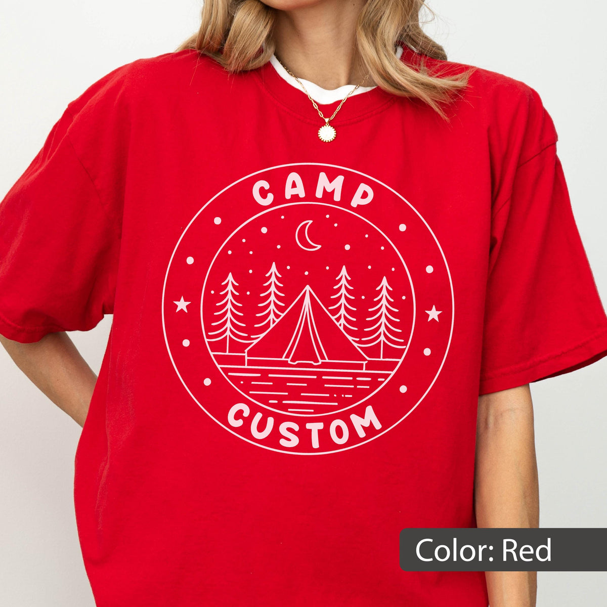 May include: A bright red short-sleeved t-shirt featuring a white circular graphic. The design includes a tent, pine trees, a crescent moon, stars, and the text "CAMP CUSTOM" printed in a bold, sans-serif font within the circular border.