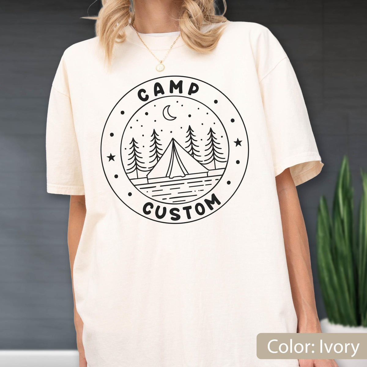 May include: An ivory short-sleeve t-shirt featuring a black circular graphic on the chest. The design includes a tent, pine trees, a crescent moon, and stars, with the text CAMP CUSTOM printed in a bold, rounded font along the circular border.