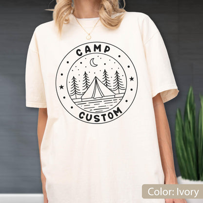 May include: An ivory short-sleeve t-shirt featuring a black circular graphic on the chest. The design includes a tent, pine trees, a crescent moon, and stars, with the text CAMP CUSTOM printed in a bold, rounded font along the circular border.
