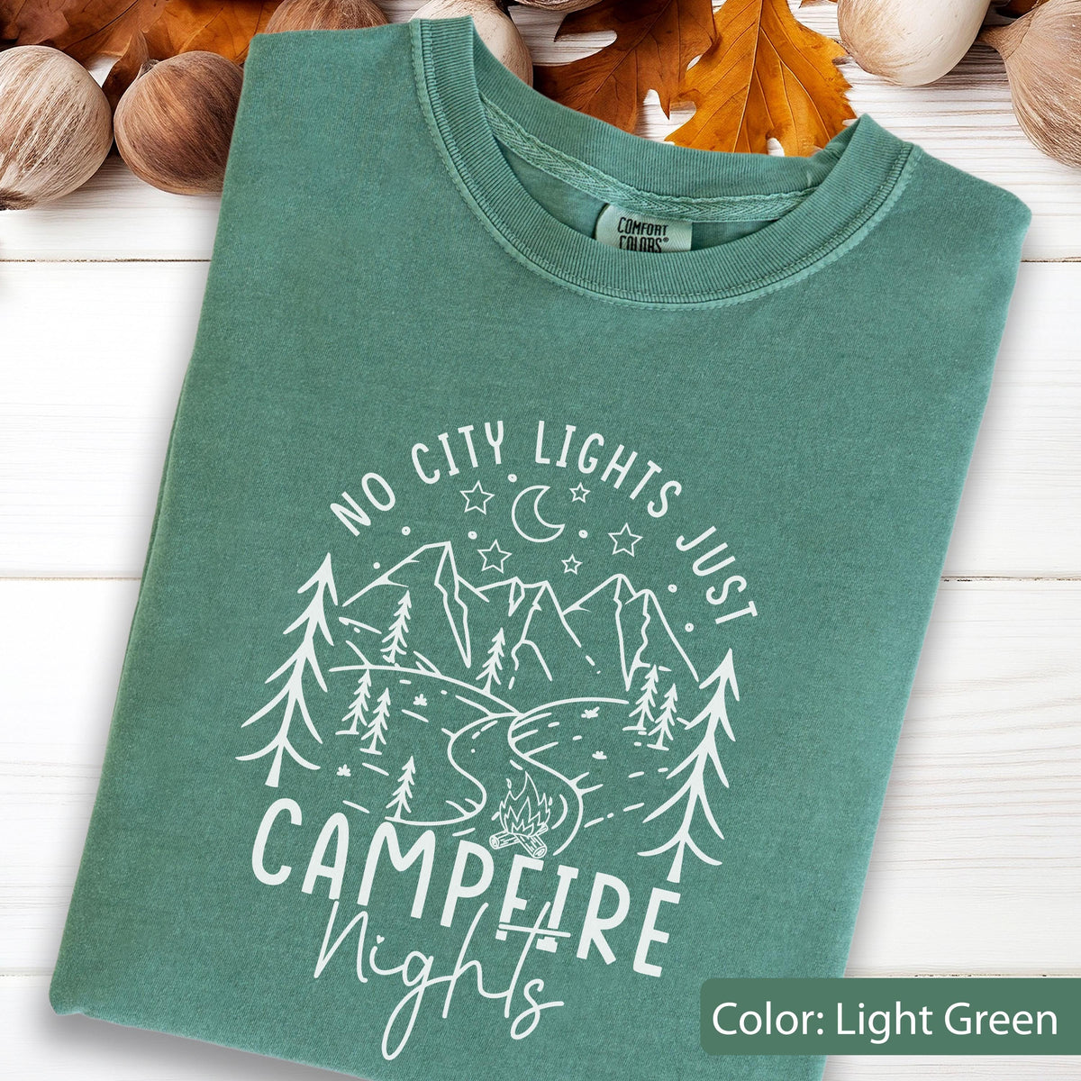 May include: A light green t-shirt featuring a white graphic print of mountains, pine trees, a winding river, a campfire, stars, and a crescent moon. The text reads NO CITY LIGHTS JUST CAMPFIRE NIGHTS. The shirt has a crew neckline and short sleeves.