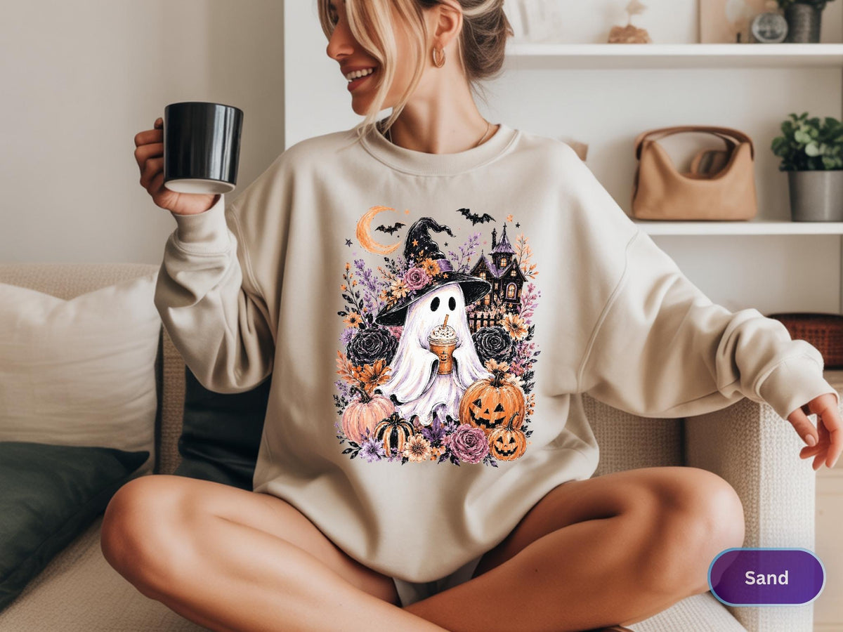 May include: A sand-colored crewneck sweatshirt featuring a graphic print of a ghost wearing a witch hat and holding a coffee cup, surrounded by pumpkins, flowers, bats, a crescent moon, and a haunted house illustration on the front.