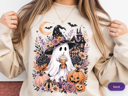 May include: A sand-colored crewneck sweatshirt featuring a graphic print of a ghost wearing a witch hat and holding a coffee cup. The design includes a crescent moon, bats, a haunted house, pumpkins, and various floral elements in orange and purple.