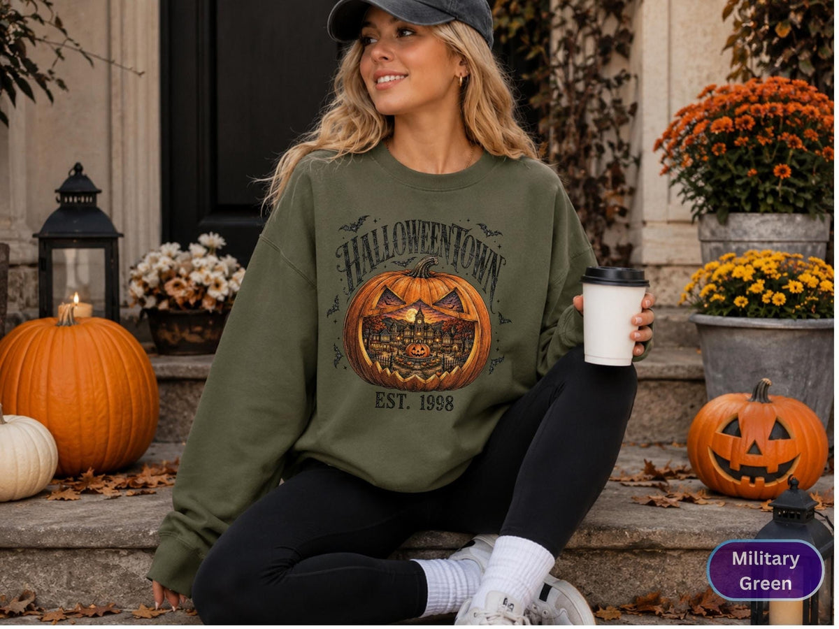 May include: A military green crewneck sweatshirt featuring a large graphic of a carved jack-o'-lantern with a town scene inside. The text reads HALLOWEENTOWN and EST. 1998 in a dark font. The sweatshirt has long sleeves and a ribbed collar.