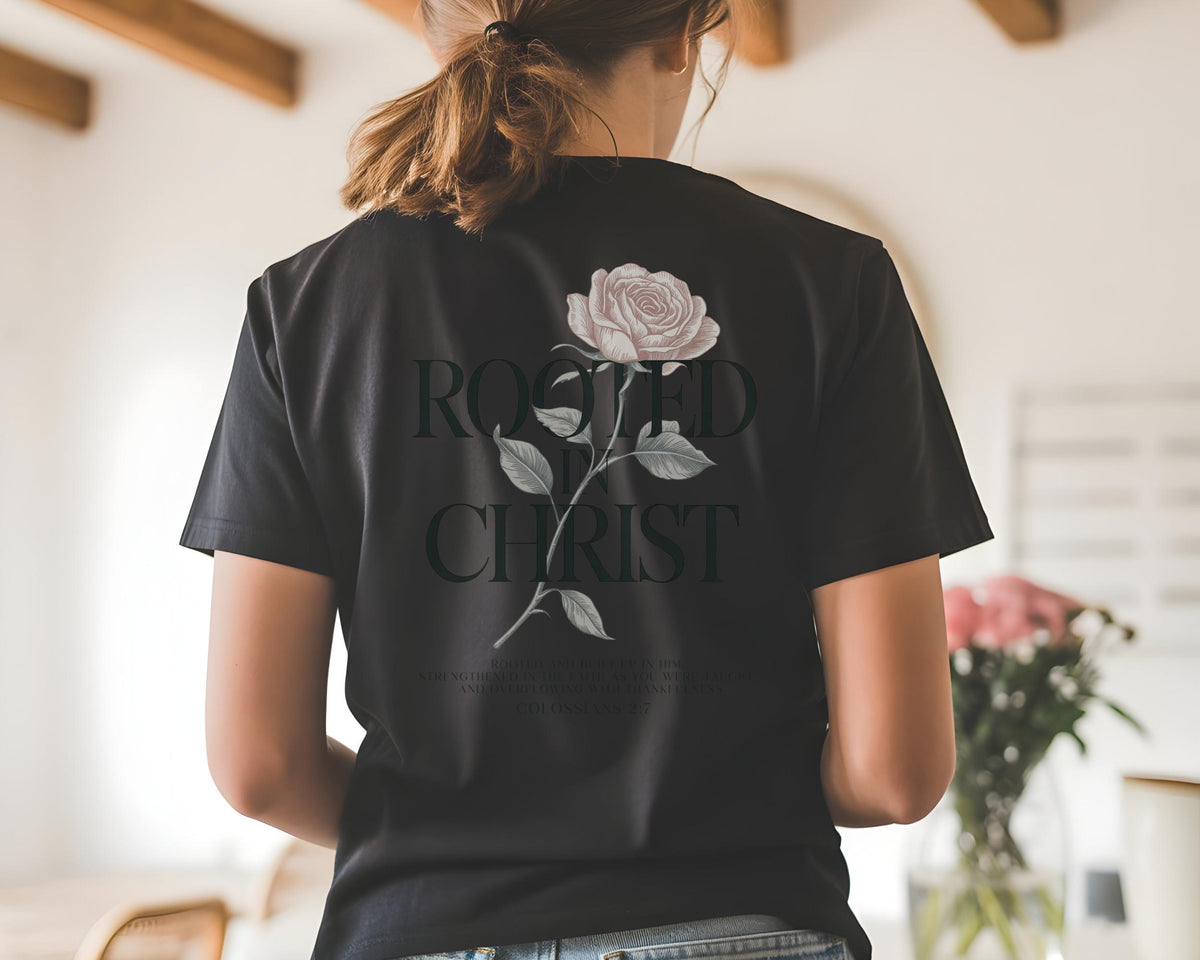 May include: A black short-sleeve t-shirt featuring a graphic on the back. The design includes a pale pink rose with green leaves and the black text "ROOTED IN CHRIST" printed in a serif font. The shirt has a crew neckline and a standard fit.
