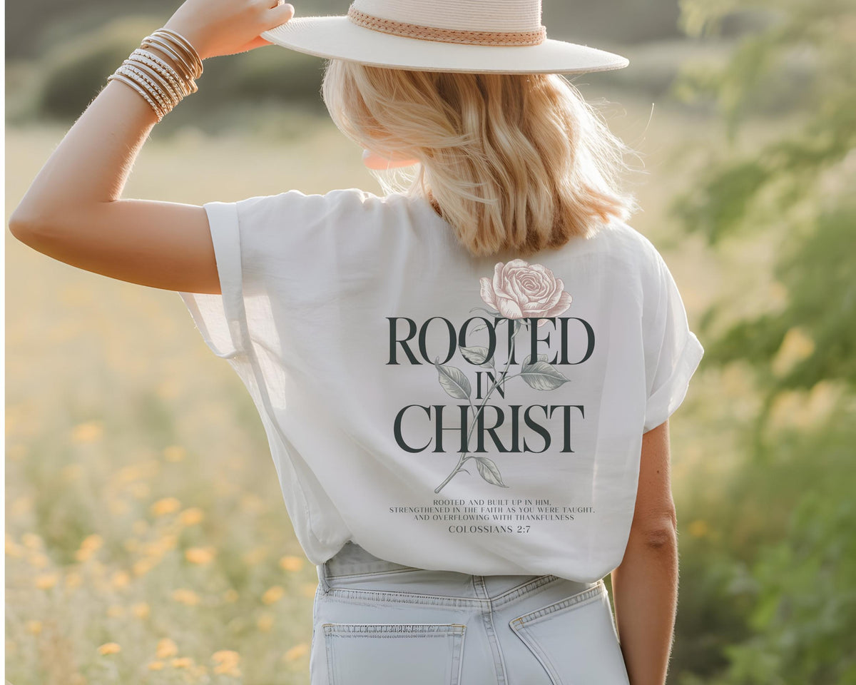 May include: A white short-sleeved t-shirt featuring a graphic on the back with a pink rose illustration, the text ROOTED IN CHRIST, and smaller text reading ROOTED AND BUILT UP IN HIM, STRENGTHENED IN THE FAITH AS YOU WERE TAUGHT, AND OVERFLOWING WITH THANKFULNESS, COLOSSIANS 2:7.