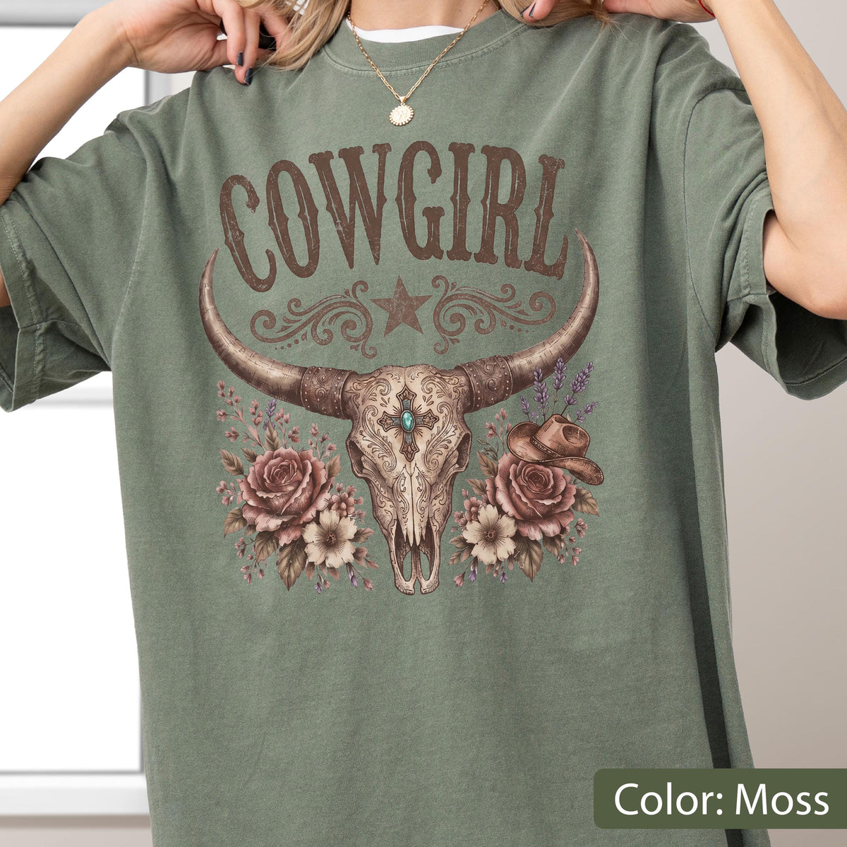 May include: A moss green t-shirt featuring a graphic print of a cow skull with long horns, a central blue stone, and intricate filigree patterns. The design includes brown roses, a small cowboy hat, and the text COWGIRL in a brown serif font.