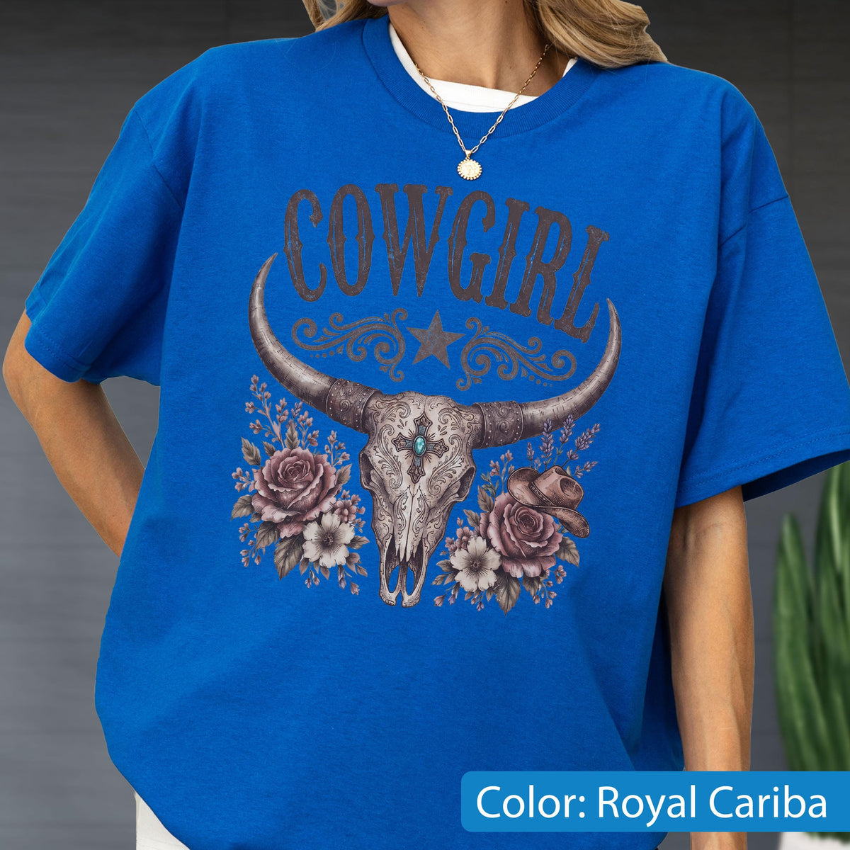 May include: A royal blue short-sleeve t-shirt featuring a graphic print of a steer skull with long horns, centered below the text COWGIRL. The design includes a central star, decorative scrollwork, two large roses, and a small cowboy hat.