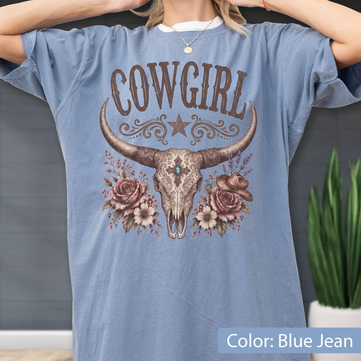 May include: A blue jean colored t-shirt featuring a graphic print of a longhorn skull, roses, and a small hat. The text COWGIRL is printed in brown above the skull. The design includes intricate floral details and a central turquoise stone accent.