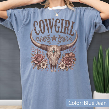 May include: A blue jean colored t-shirt featuring a graphic print of a longhorn skull, roses, and a small hat. The text COWGIRL is printed in brown above the skull. The design includes intricate floral details and a central turquoise stone accent.