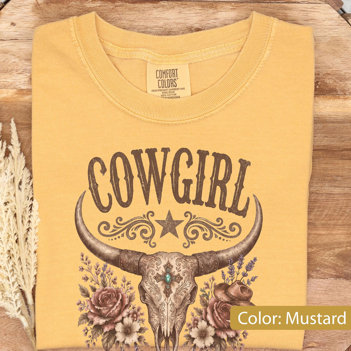 May include: A mustard yellow t-shirt featuring a brown graphic of a cow skull with long horns, centered above a floral arrangement of roses and a small hat. The text COWGIRL is printed in a brown western-style font above the skull design.