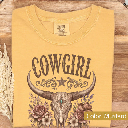 May include: A mustard yellow t-shirt featuring a brown graphic of a cow skull with long horns, centered above a floral arrangement of roses and a small hat. The text COWGIRL is printed in a brown western-style font above the skull design.