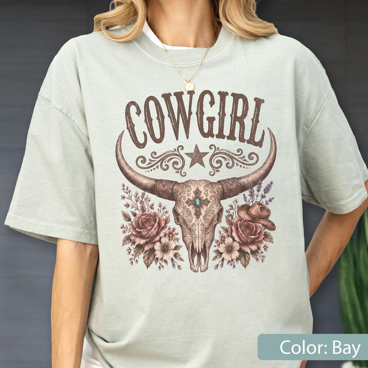 May include: A light sage green short-sleeve t-shirt featuring a brown graphic print of a longhorn skull, roses, a cowboy hat, and the text COWGIRL. The design includes decorative scrollwork, a star, and a central turquoise-colored accent.