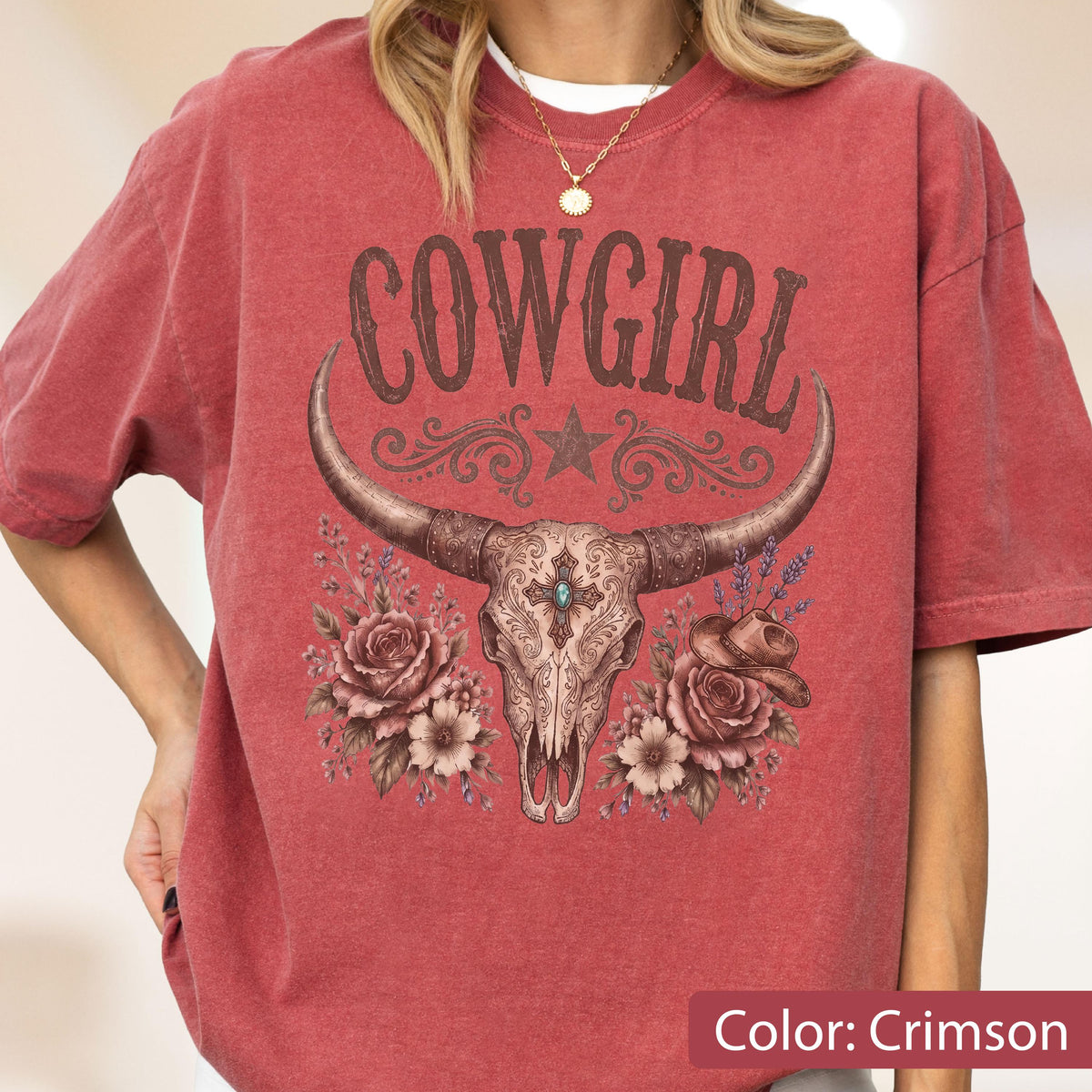 May include: A crimson short-sleeve t-shirt featuring a graphic print of a cow skull with long horns, centered above a cluster of roses and a small cowboy hat. The text COWGIRL is printed in a dark brown font above the skull, with a star icon.