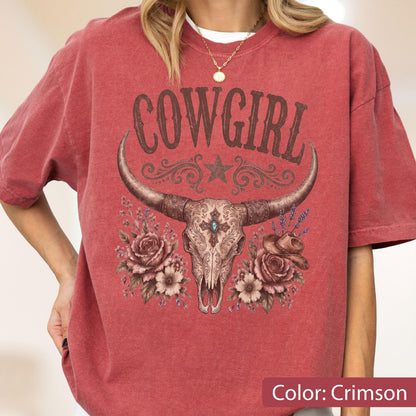 May include: A crimson short-sleeve t-shirt featuring a graphic print of a cow skull with long horns, centered above a cluster of roses and a small cowboy hat. The text COWGIRL is printed in a dark brown font above the skull, with a star icon.