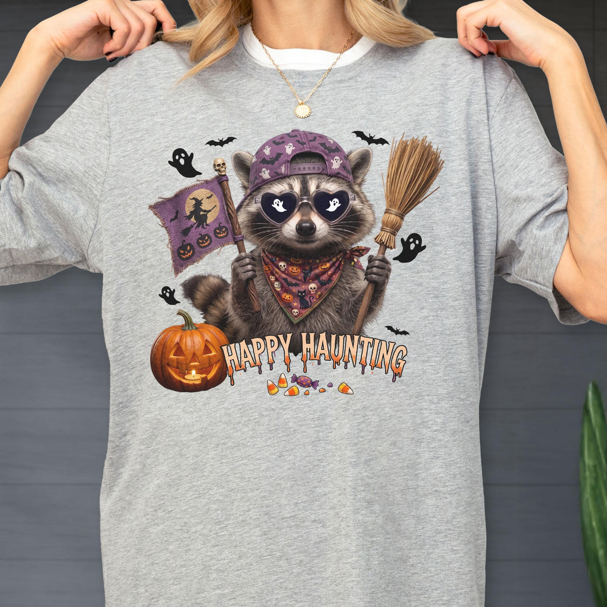 May include: A heather gray t-shirt featuring a graphic of a raccoon wearing a purple baseball cap, heart-shaped sunglasses with ghost silhouettes, and a patterned bandana. The raccoon holds a broom and a purple flag with witch and pumpkin icons.