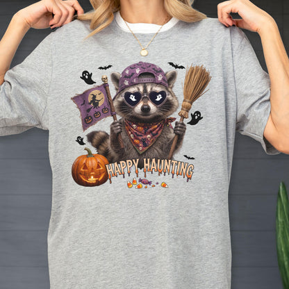 May include: A heather gray t-shirt featuring a graphic of a raccoon wearing a purple baseball cap, heart-shaped sunglasses with ghost silhouettes, and a patterned bandana. The raccoon holds a broom and a purple flag with witch and pumpkin icons.