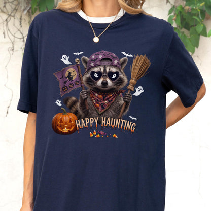 May include: A navy blue t-shirt featuring a graphic of a raccoon wearing a purple cap, heart-shaped sunglasses, and a patterned bandana. The raccoon holds a broom and a flag with a witch silhouette. Text reads HAPPY HAUNTING with pumpkins and ghosts.