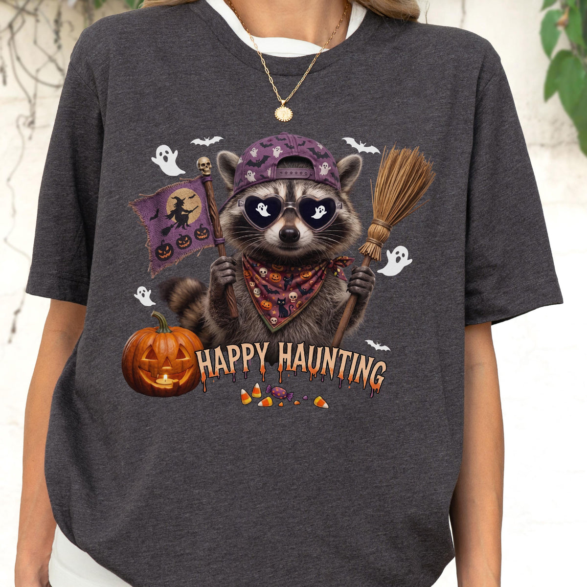 May include: A charcoal gray t-shirt featuring a graphic of a raccoon wearing a purple baseball cap, heart-shaped sunglasses, and a patterned bandana. The raccoon holds a broom and a flag with a witch. Text reads HAPPY HAUNTING with pumpkins.
