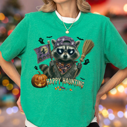 May include: A bright green t-shirt featuring a graphic of a raccoon wearing a baseball cap and heart-shaped sunglasses. The raccoon holds a broom and a flag with a witch silhouette. The text HAPPY HAUNTING is printed below with pumpkins and candy.