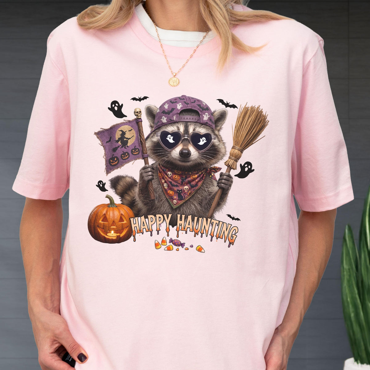 May include: A light pink t-shirt featuring a graphic of a raccoon wearing a purple baseball cap, heart-shaped sunglasses, and a patterned bandana. The raccoon holds a broom and a flag with a witch silhouette. Text reads Happy Haunting.
