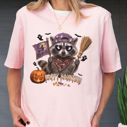 May include: A light pink t-shirt featuring a graphic of a raccoon wearing a purple baseball cap, heart-shaped sunglasses, and a patterned bandana. The raccoon holds a broom and a flag with a witch silhouette. Text reads Happy Haunting.