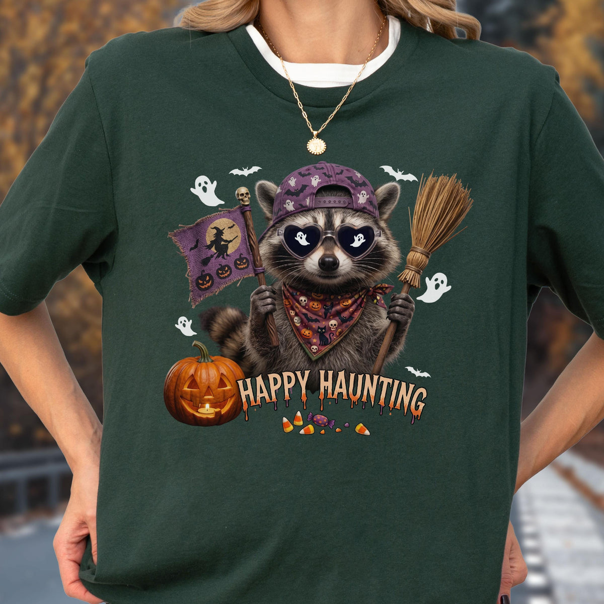 May include: A dark green t-shirt featuring a graphic of a raccoon wearing a purple baseball cap and heart-shaped sunglasses. The raccoon holds a flag with a witch and a broomstick. Text reads HAPPY HAUNTING above candy corn and a jack-o'-lantern.