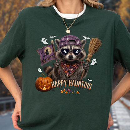 May include: A dark green t-shirt featuring a graphic of a raccoon wearing a purple baseball cap and heart-shaped sunglasses. The raccoon holds a flag with a witch and a broomstick. Text reads HAPPY HAUNTING above candy corn and a jack-o'-lantern.