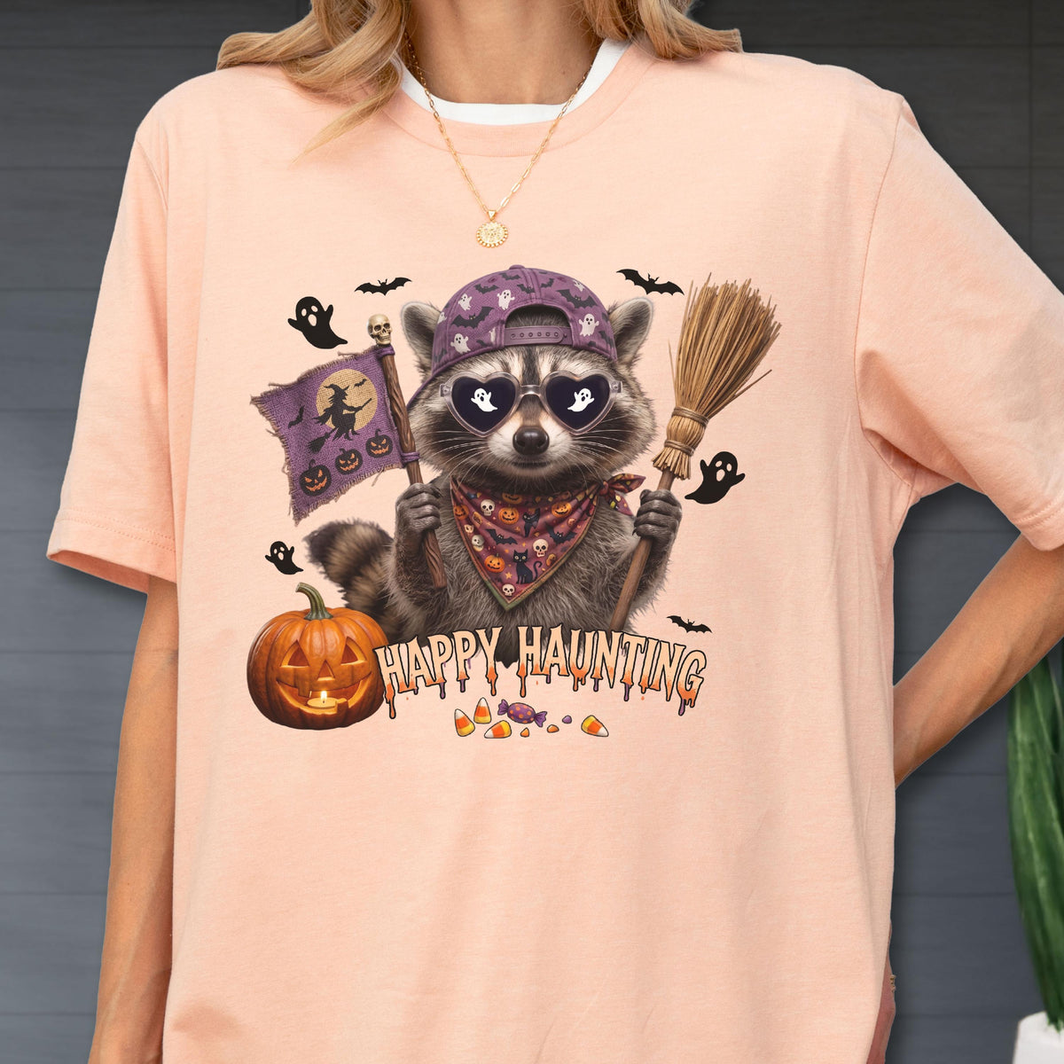 May include: A peach-colored t-shirt featuring a graphic of a raccoon wearing a purple baseball cap, heart-shaped sunglasses, and a patterned bandana. The raccoon holds a broom and a flag with a witch silhouette. Text reads HAPPY HAUNTING.