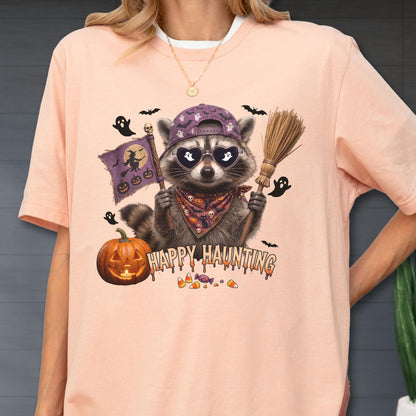 May include: A peach-colored t-shirt featuring a graphic of a raccoon wearing a purple baseball cap, heart-shaped sunglasses, and a patterned bandana. The raccoon holds a broom and a flag with a witch silhouette. Text reads HAPPY HAUNTING.