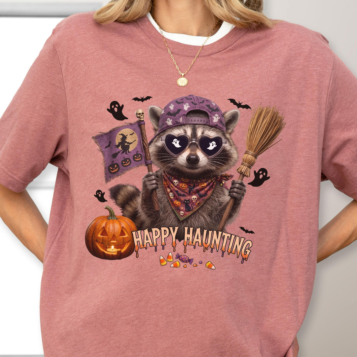 May include: A mauve t-shirt featuring a graphic of a raccoon wearing a purple baseball cap, heart-shaped sunglasses, and a patterned bandana. The raccoon holds a broom and a flag with a witch silhouette. Text reads Happy Haunting.
