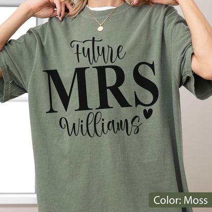 May include: A moss green short-sleeved t-shirt featuring black printed text that reads Future MRS Williams with a small heart icon. The shirt has a crew neckline and a relaxed fit, showcasing a bold graphic design centered on the chest area.
