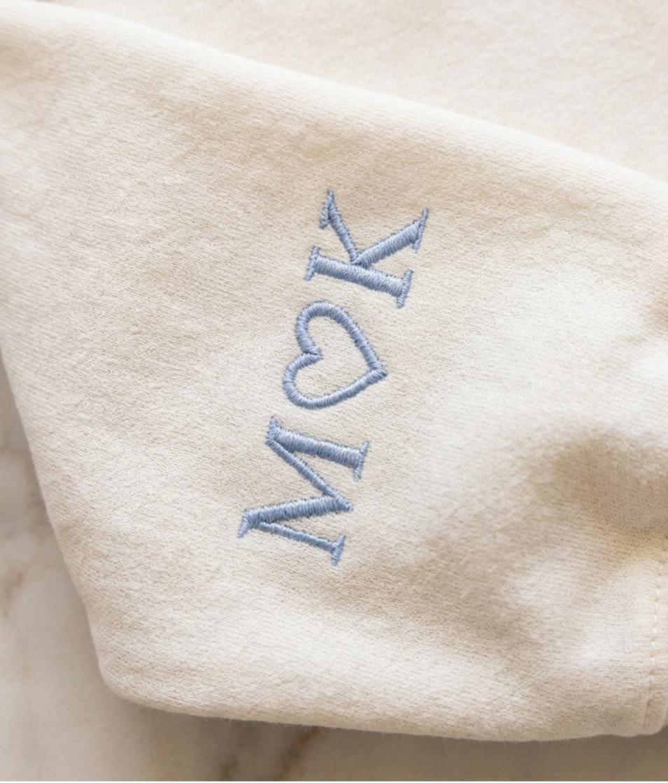 May include: A cream-colored fabric featuring light blue embroidery. The stitched design consists of the letter M, a heart shape, and the letter K arranged vertically. The embroidery uses a classic serif font style with clean, precise thread work.
