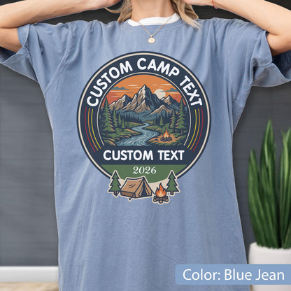 May include: A blue jean colored t-shirt featuring a circular graphic with a mountain landscape, a river, and a campfire. The text reads CUSTOM CAMP TEXT at the top and bottom, with the year 2026 printed below. A small tent and pine trees are visible.
