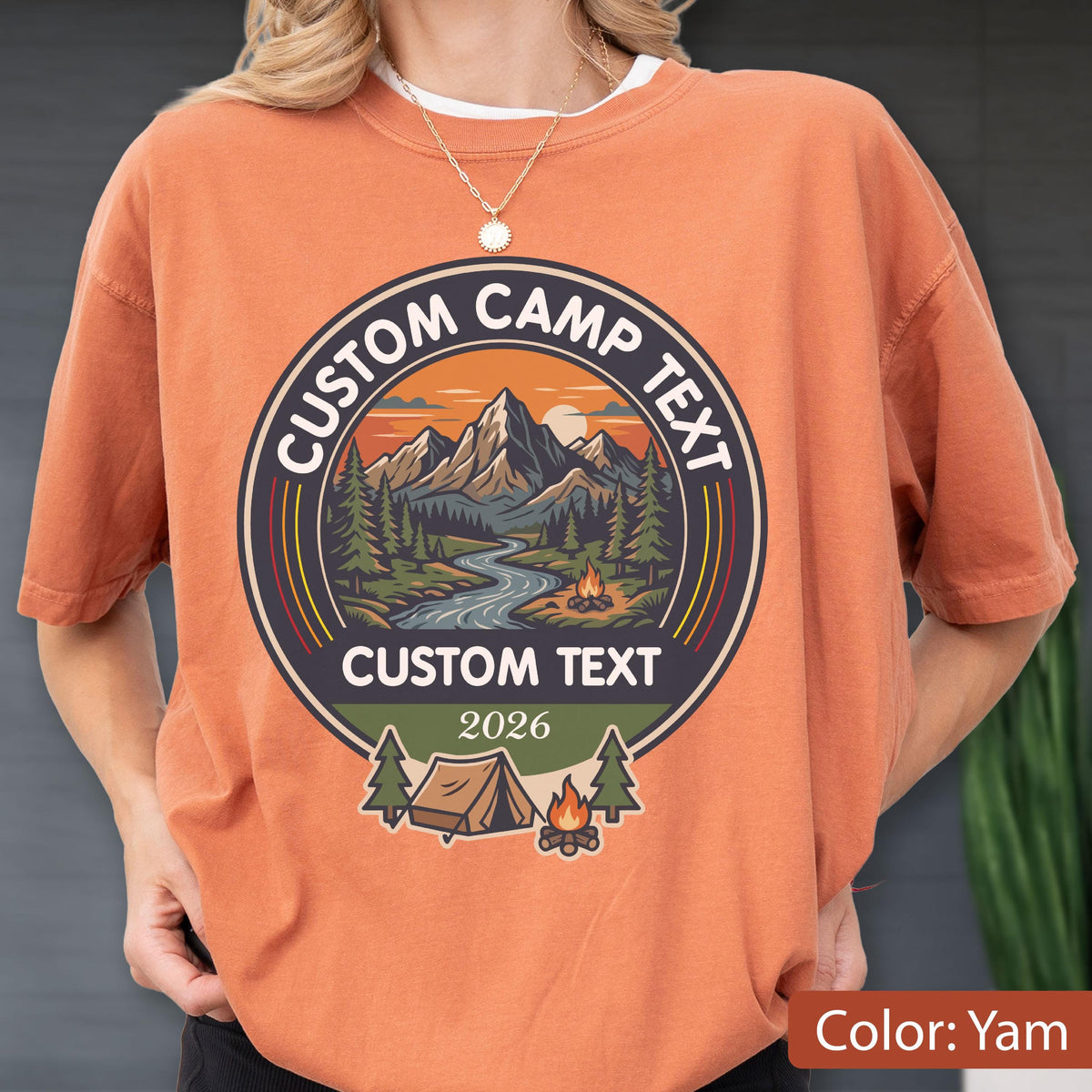 May include: A yam-colored t-shirt featuring a circular graphic with a mountain landscape, a river, and a campfire. The text reads CUSTOM CAMP TEXT at the top and bottom, with the year 2026 below. A tent and pine trees are depicted at the base.