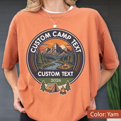 May include: A yam-colored t-shirt featuring a circular graphic with a mountain landscape, a river, and a campfire. The text reads CUSTOM CAMP TEXT at the top and bottom, with the year 2026 below. A tent and pine trees are depicted at the base.