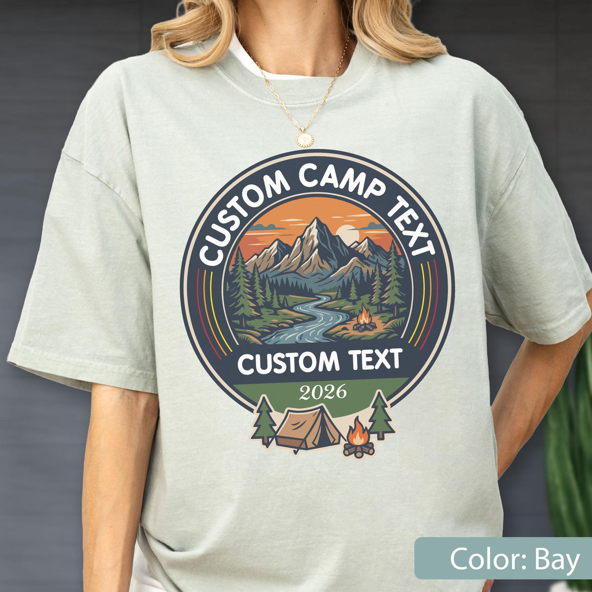 May include: A light sage green t-shirt featuring a circular graphic with a mountain landscape, a river, a campfire, and a tent. The design includes the text CUSTOM CAMP TEXT at the top and bottom, with the year 2026 printed in the center of the graphic.