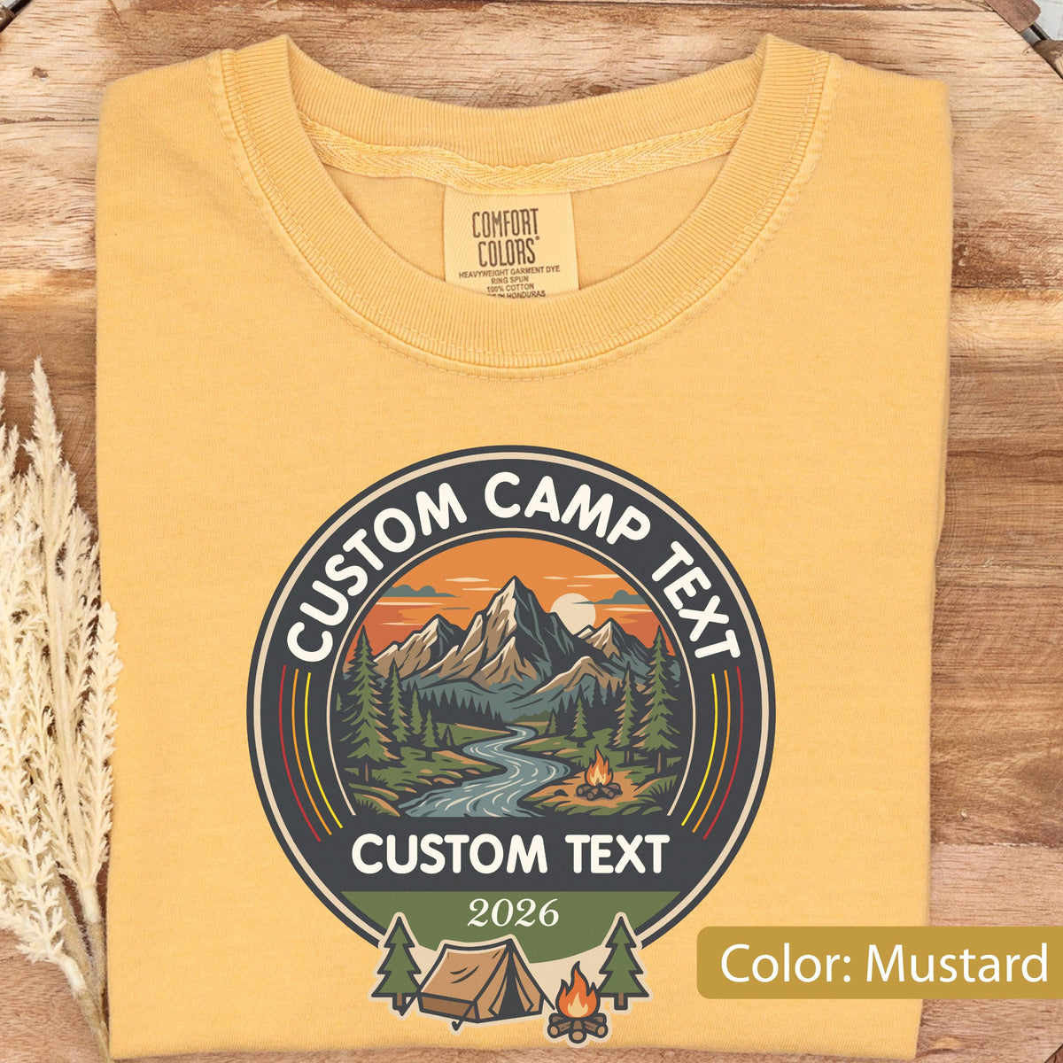May include: A mustard yellow t-shirt featuring a circular graphic with a mountain landscape, a river, a campfire, and a tent. The text reads CUSTOM CAMP TEXT, CUSTOM TEXT, and 2026. The shirt has a Comfort Colors label inside the crew neckline.