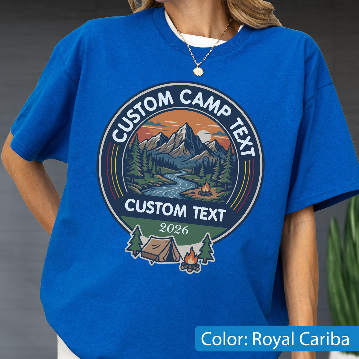 May include: A royal blue t-shirt featuring a circular graphic with a mountain landscape, a river, and a campfire. The text reads CUSTOM CAMP TEXT at the top and bottom, with the year 2026 below. Below the circle is a tent and a small campfire icon.