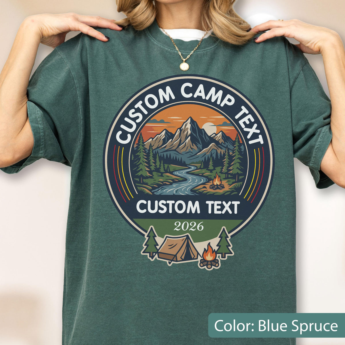 May include: A blue spruce t-shirt featuring a circular graphic with a mountain landscape, a river, a campfire, and a tent. The design includes the text CUSTOM CAMP TEXT, CUSTOM TEXT, and the year 2026 in a retro-inspired color palette.