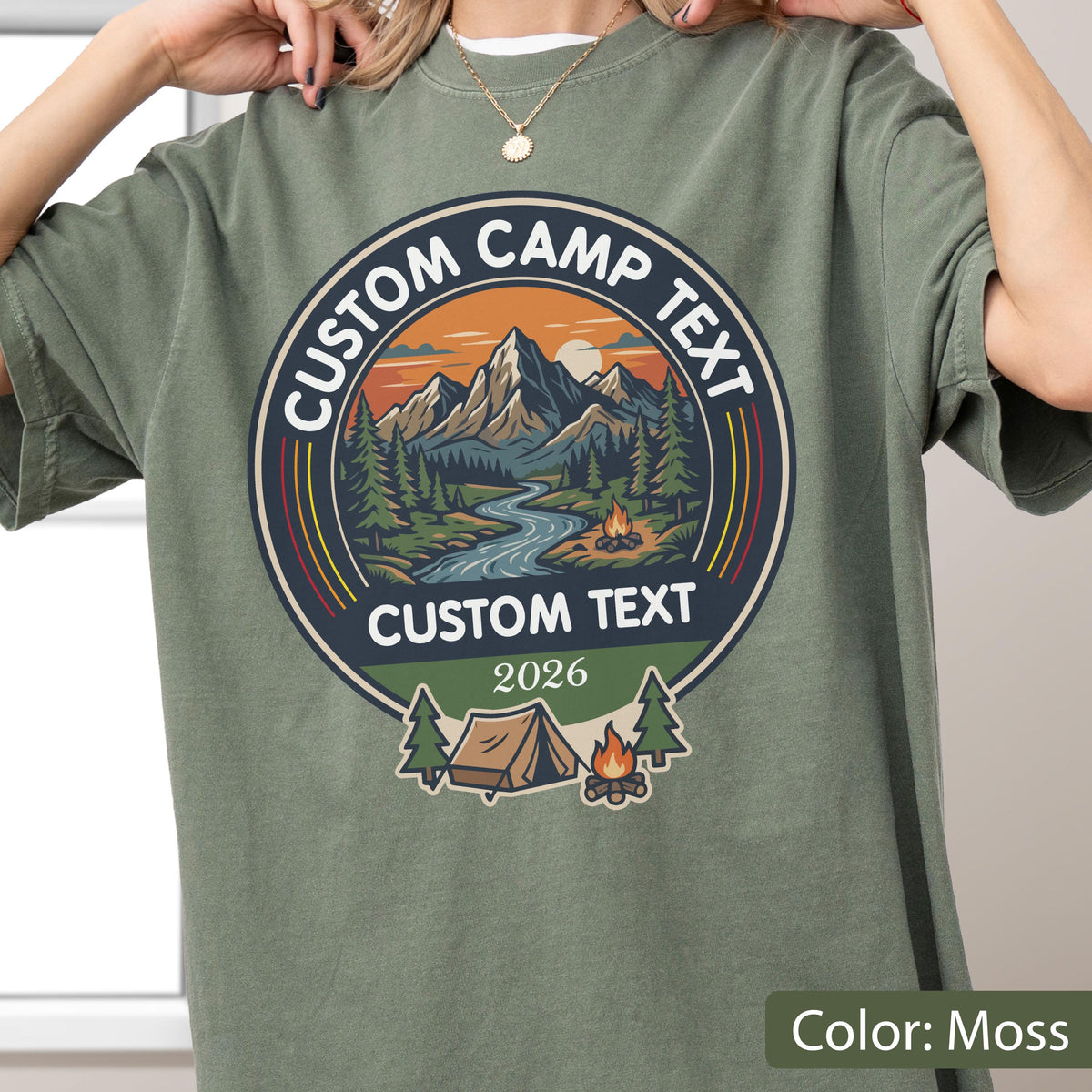 May include: A moss green t-shirt featuring a circular graphic with a mountain landscape, a river, and a campfire. The text reads CUSTOM CAMP TEXT, CUSTOM TEXT, and 2026. Below the circle is a small tent and another campfire illustration.