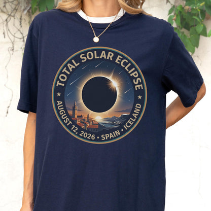 May include: A navy blue t-shirt featuring a circular graphic of a total solar eclipse over a coastal town. The text reads TOTAL SOLAR ECLIPSE AUGUST 12, 2026 SPAIN ICELAND. The design includes a dark sun, a bright corona, and a starry night sky.