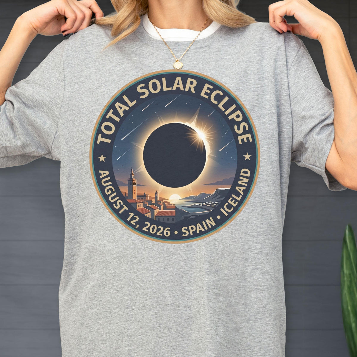 May include: A heather gray t-shirt featuring a circular graphic of a total solar eclipse over a coastal town. The text reads Total Solar Eclipse, August 12, 2026, Spain, Iceland. The design includes a dark moon, a glowing sun corona, and stars.